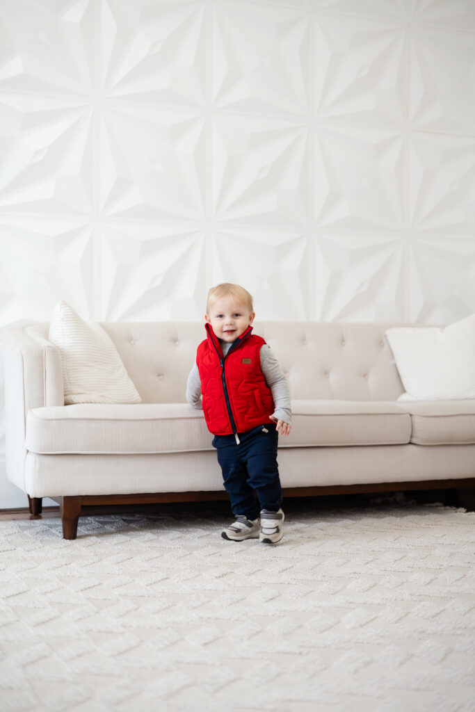 Toddler boy standing by a couch with a sweet smile on his face